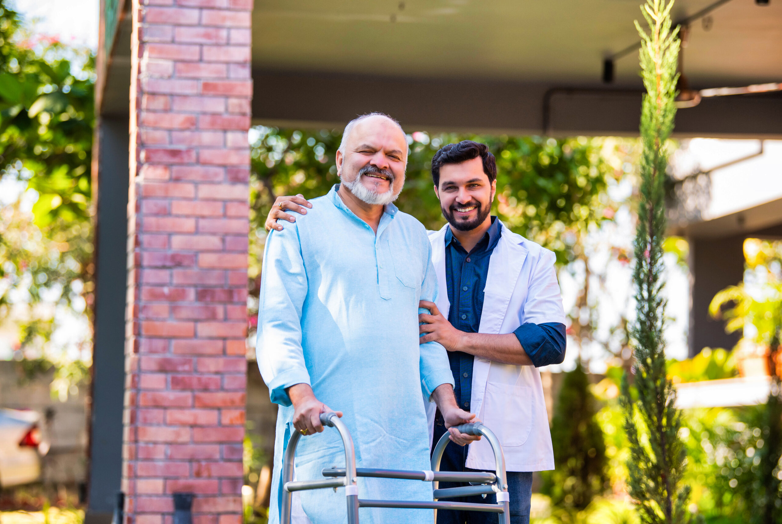 Indian Asian handsome male doctor accompanies senior old-aged male patient using a walker in a hospital lawn garden, engaging in a thoughtful discussion about the medical case and healthcare recovery