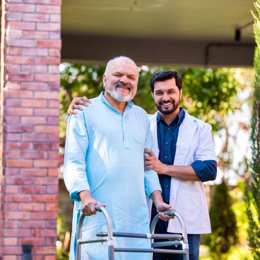 Indian Asian handsome male doctor accompanies senior old-aged male patient using a walker in a hospital lawn garden, engaging in a thoughtful discussion about the medical case and healthcare recovery